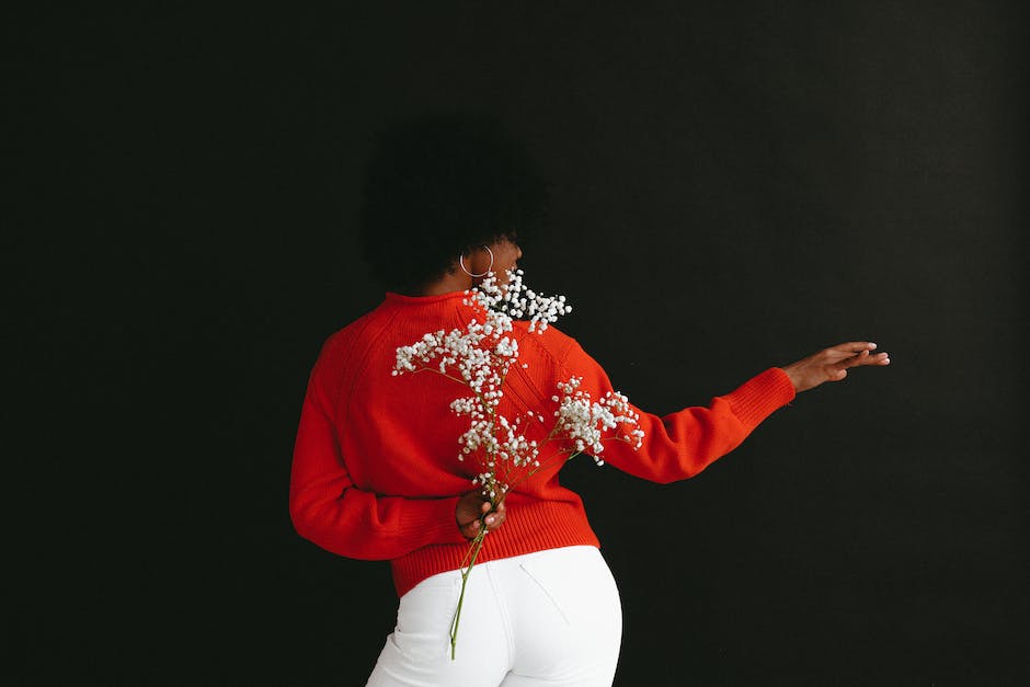 Back view of sensual black woman in white denim and white red sweater holding Gypsophila flower behind back posing on black backdrop